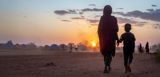 Silhuette of a woman and child hand in hand walking on sand during a sunset.